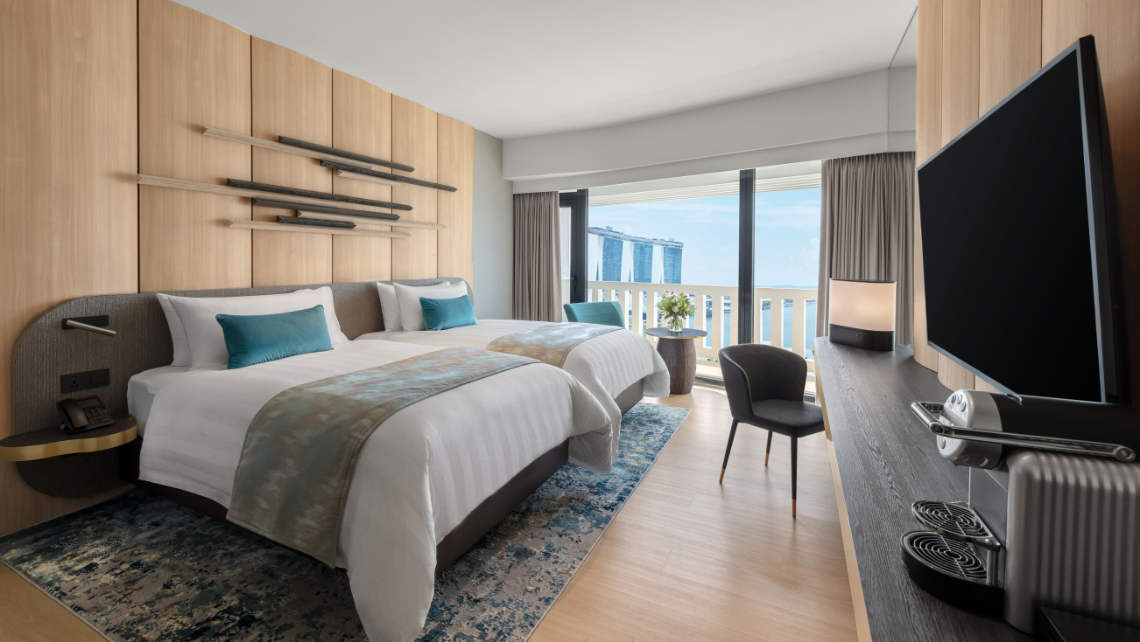A modern hotel room with two beds, a large window offering panoramic views of the Singapore city skyline, and a flat-screen TV.
