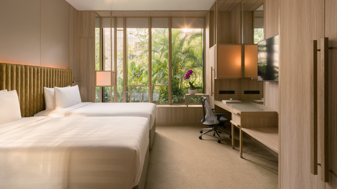 A bright hotel room with two beds, a desk area, and large windows looking out onto lush greenery, reflecting a garden-in-a-hotel concept.