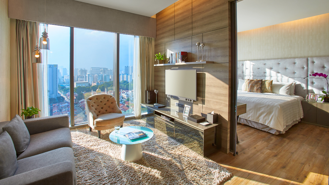 A spacious and well-lit hotel suite with a living area featuring a grey sofa and armchair, and a separate bedroom area with a large bed, all with city views from expansive windows.